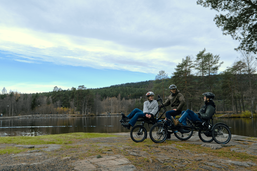 2 personer på en blå tandemsykkel og 1 person i liggesykkel foran et vann med skog bak, alle smiler til hverandre